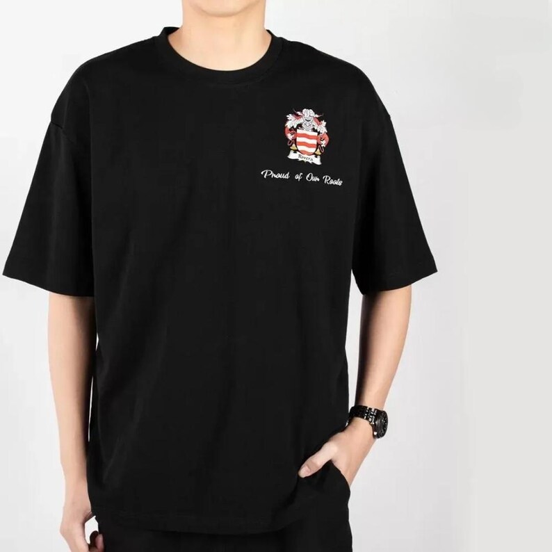 Może przedstawiać: Czarny T-shirt z kr&oacute;tkim rękawem, z haftowanym białym herbem i napisem "Proud of Our Roots".