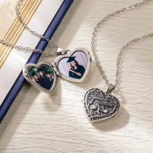 May include: Silver heart-shaped locket necklaces. One locket is open, revealing a photo of a graduate in a cap and gown. The other locket is closed, with a graduation cap design. Both are on delicate chains.