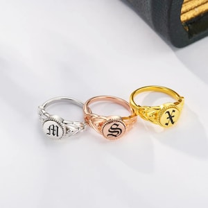 Könnte beinhalten: Drei Siegelringe in Silber, Roségold und Gold. Jeder Ring hat einen anderen Anfangsbuchstaben: "M", "S" und "X". Die Ringe haben ein dekoratives Band und eine kreisförmige Oberfläche mit dem schwarzen Anfangsbuchstaben.