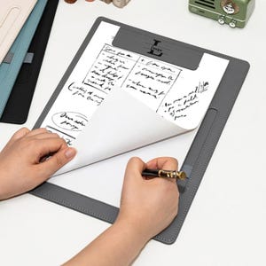 May include: A gray clipboard with a pen and handwritten notes. The clipboard has a leather-like texture and a metal clip at the top. The pen is black with gold accents. The notes are in black ink.