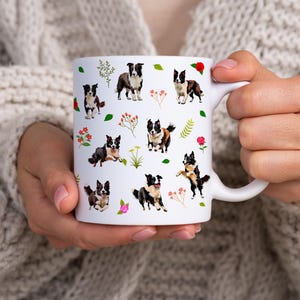 May include: White ceramic mug featuring a repeating pattern of Border Collie dogs in various poses, interspersed with floral and leaf accents. The dogs are depicted in black, white, and brown. The mug is held by a person wearing a cream-colored sweater.