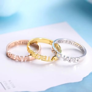 May include: Three rings in rose gold, gold, and silver. The rose gold ring spells "DREAM", the gold ring spells "&DELLA", and the silver ring spells "HARD". The rings are on a white surface.
