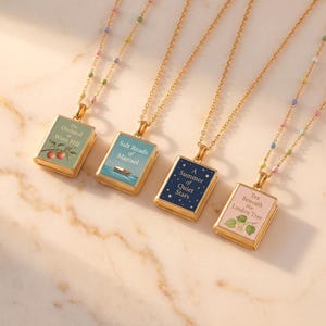 May include: Four gold-toned necklaces featuring rectangular book-shaped pendants. The pendants display titles: The Orchard at Wren Hill, Salt Roads of Marisol, A Summer of Quiet Stars, and Tea Beneath the Linden Tree. The chains feature small colorful beads.