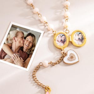 Op de afbeelding: Een goudkleurige ketting met een medaillon met twee kleine foto's. De ketting heeft een ketting met een hartvormige hanger en een rij roze kralen. Links staat een foto van twee vrouwen.