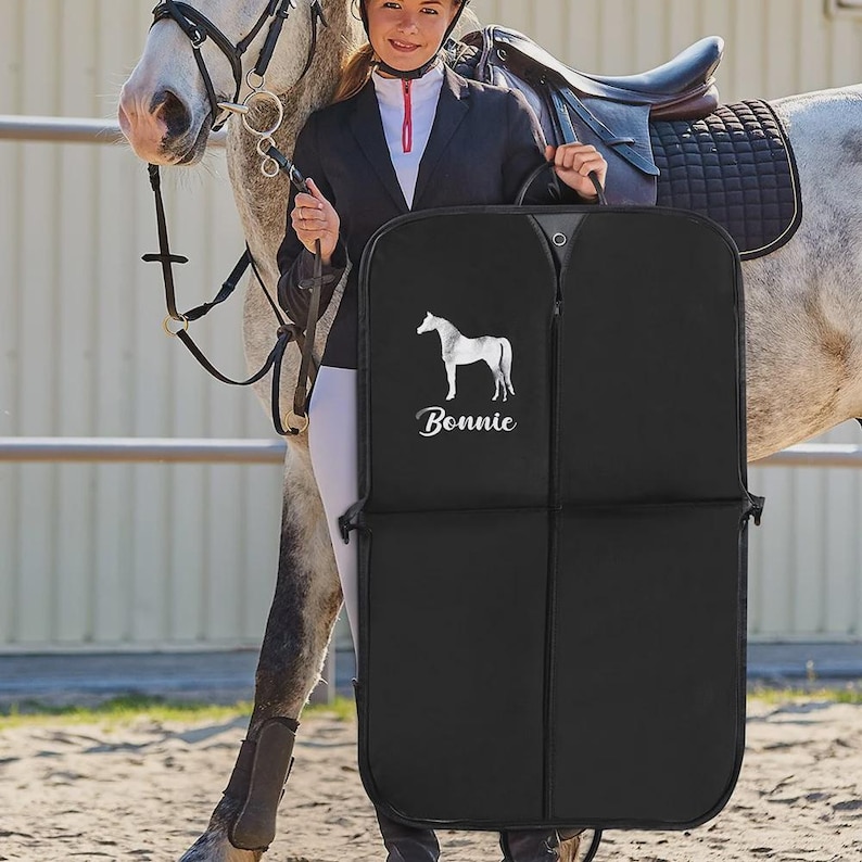 Puede incluir: Bolsa de ropa negra con una silueta de caballo blanca y el nombre "Bonnie" impreso. La bolsa la sostiene una persona con atuendo ecuestre, de pie junto a un caballo gris ensillado.