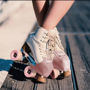 Puede incluir: Patines de ruedas color crema con ruedas y detalles rosas. Los patines tienen un diseño de caña alta con cordones y una puntera rosa con la palabra "love" en escritura blanca. Los patines están sobre una superficie de madera.