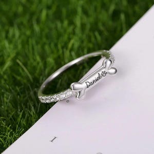 May include: A silver ring featuring a bone-shaped charm inscribed with the name "Daniel" and a paw print. The ring's band is set with small, clear stones. The ring rests on a white surface, with green grass visible in the background.