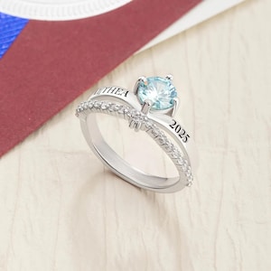 May include: A silver ring with a large, light blue gemstone at the center. The band is split, with one side inscribed with "THEA" and the other with "2025". Small, clear stones line the split band, adding sparkle.