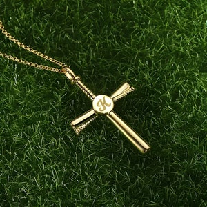 Peut inclure: Un collier avec un pendentif en forme de croix dorée et une chaîne délicate. La croix porte la lettre "K" gravée dans un cercle au centre. Le collier est présenté sur une surface herbeuse verte.