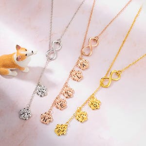 May include: Three gold necklaces with infinity symbols and dangling charms in the shape of paw prints with letters on them. The charms spell out the names 'ALS', 'LEG', and 'SGJ'.