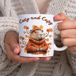 Könnte beinhalten: Weiße Keramik-Tasse mit den Worten "Cozy and Capy" in brauner Schrift. Die Tasse zeigt ein Capybara mit orangefarbener Mütze und Pullover, umgeben von Herbstblättern. Die Tasse wird von einer Person gehalten, die einen cremefarbenen Pullover trägt.