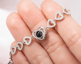 May include: A silver bracelet with a heart-shaped charm that projects a message when held up to a light. The bracelet is made of small, clear cubic zirconia stones and features a chain of connected hearts.