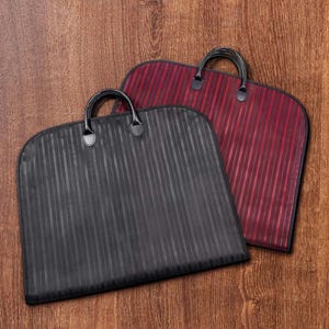 Puede incluir: Dos fundas para ropa con asas. Una es negra con rayas verticales, la otra es burdeos con rayas verticales. Ambas bolsas tienen asas negras y est&aacute;n sobre una superficie de madera.
