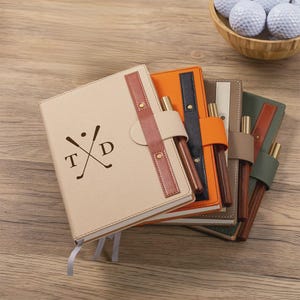 May include: A collection of personalized golf-themed notebooks in various colors, including beige, orange, navy, and green. Each notebook features a pen holder and a golf club and ball design with initials. A wooden bowl of golf balls is in the background.