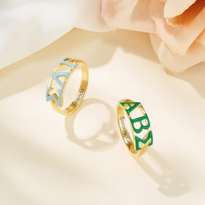 Könnte beinhalten: Zwei goldene Ringe mit griechischen Buchstaben aus hellblauem und grünem Emaille. Ein Ring hat den eingravierten Namen "Brenda", der andere sagt "Sorority Girl". Die Ringe befinden sich auf hellem Hintergrund.