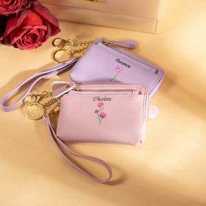 May include: Two small, zippered coin purses in shades of lavender and pink, with gold-tone hardware and wrist straps. Each purse features an embroidered floral design and a name: "Florence" and "Charlotte".