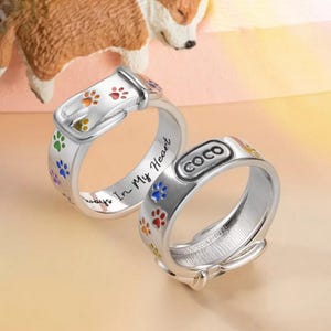Op de afbeelding: Twee zilveren ringen met een ontwerp van een hondenhalsband. Elke ring heeft kleurrijke pootafdrukken en een gespdetail. Eén ring heeft de naam "COCO" en de inscriptie "Always In My Heart".