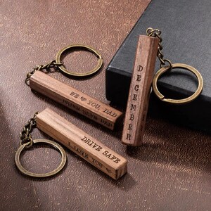 May include: Three wooden keychains with bronze-colored key rings and chains. The keychains are engraved with the phrases "WE ❤️ YOU, DAD", "DRIVE SAFE I LOVE YOU", and "DECEMBER". The keychains are rectangular and made of a light brown wood.
