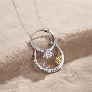 May include: A silver-toned necklace featuring two interlocking circular pendants. The top circle holds a clear, faceted stone setting. The bottom circle displays a small gold-toned sun, a silver crescent moon, and the engraved text L+T 08.17.25.