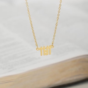 May include: A gold-colored necklace with the numbers "11:11" as a pendant. The necklace is on a chain and is displayed against a blurred background. The pendant is a stylized font.