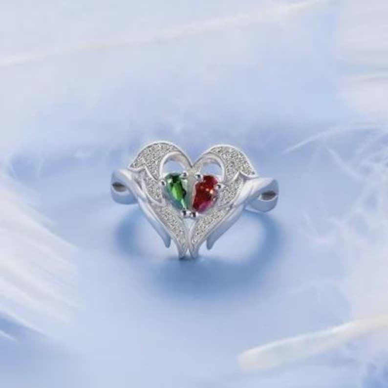 Puede incluir: Un anillo de plata con un dise&ntilde;o en forma de coraz&oacute;n. El anillo presenta dos piedras preciosas en forma de pera, una verde y otra roja, colocadas una al lado de la otra. El coraz&oacute;n est&aacute; adornado con peque&ntilde;as piedras brillantes.