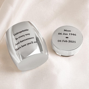 May include: A silver-colored memorial keepsake set. One piece is a faceted, curved shape with the inscription "Unforgettable In every way And forevermore That's how you'll sta". The other piece is a round container with the inscription "Mom 06 Jan 1946 04 Feb 2025".