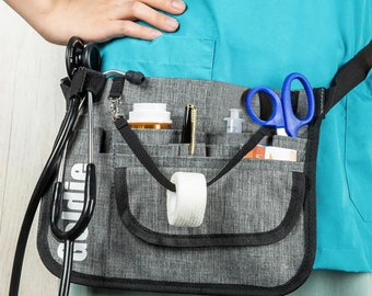 May include: A gray medical utility belt with multiple pockets and a black strap. The belt is worn over a turquoise scrub top. The belt has a stethoscope, scissors, a bottle of pills, a pen, a roll of bandage, and a small bottle of liquid in the pockets.