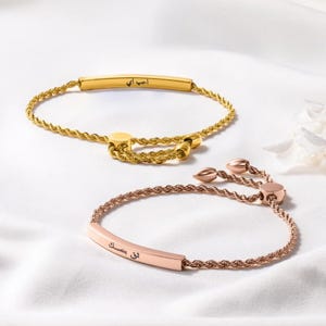 May include: Two adjustable bracelets. One is gold-toned with a rope chain and a rectangular bar with engraved text. The other is rose gold-toned, also with a rope chain and engraved bar. Both are on a white surface.