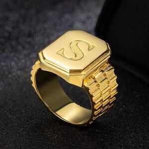 May include: A gold-toned ring with a square-shaped top featuring the letter "S" in a raised design. The band of the ring has a textured, patterned design.