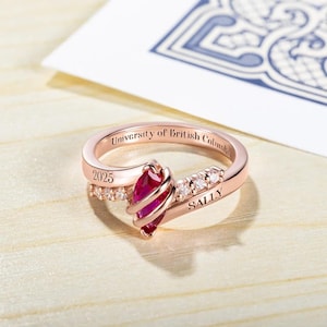 May include: A rose gold ring featuring a red gemstone and small clear gemstones. The ring is engraved with "University of British Columbia" and "Sally" along with the year "2025".