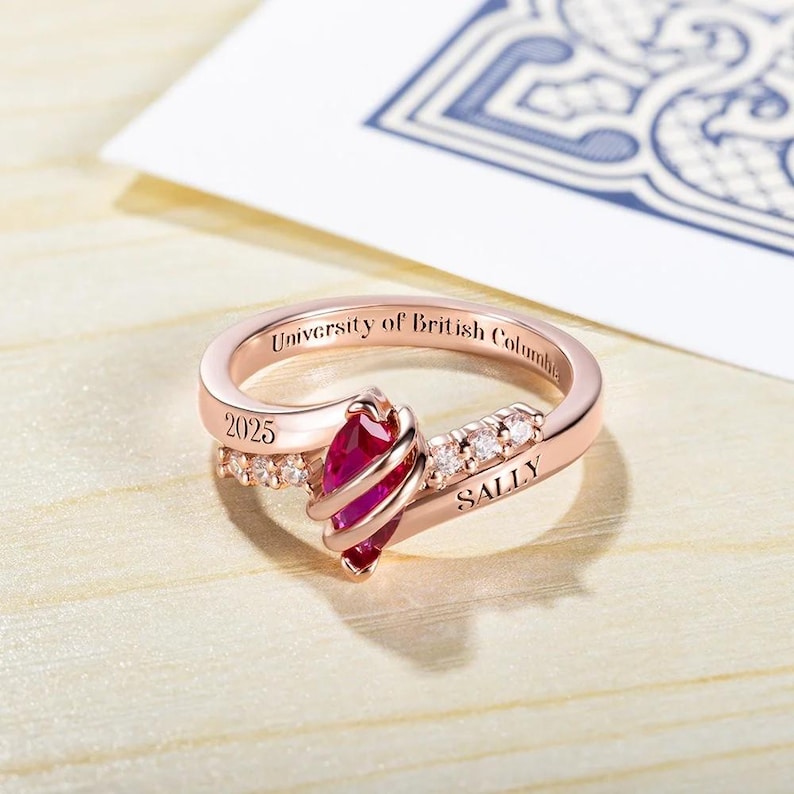 May include: A rose gold ring featuring a red gemstone and small clear gemstones. The ring is engraved with "University of British Columbia" and "Sally" along with the year "2025".