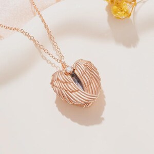 May include: Rose gold necklace featuring a pendant shaped like angel wings. The wings are detailed with layered feathers and a small, clear gemstone at the center. The chain is delicate, and the pendant casts a shadow on the light-colored surface.