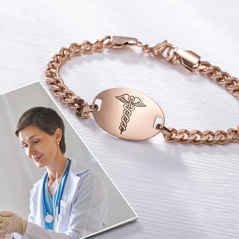 May include: A rose gold medical ID bracelet with a curb chain and an oval tag engraved with the caduceus symbol.