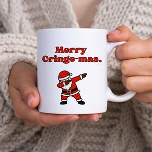 Könnte beinhalten: Weiße Keramik-Tasse mit dem Text "Merry Cringe-mas" in roten Buchstaben. Eine Cartoon-Figur des Weihnachtsmanns in rotem Anzug und Hut macht einen Dab. Die Tasse wird von einer Person gehalten, die einen cremefarbenen Pullover trägt.