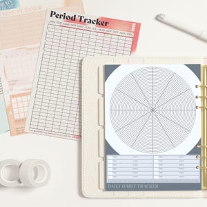 Könnte beinhalten: Ein weißer Ordner mit einer grauen Lasche mit der Aufschrift "Täglicher Gewohnheits-Tracker". Die Lasche enthält einen Kreis, der in Abschnitte unterteilt ist, wobei Linien vom Mittelpunkt ausstrahlen. Der Ordner enthält eine Kalenderseite mit der Aufschrift "Perioden-Tracker" mit den Monaten Januar bis Dezember.