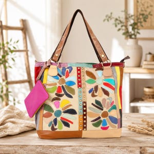 May include: A multi-colored leather tote bag featuring a patchwork design with floral motifs in shades of red, blue, green, and yellow. The bag includes a vertical strip of gold-tone metal studs, two brown leather handles, and a detachable pink pouch.