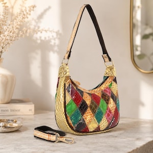 May include: A colorful patchwork shoulder bag with a cream-colored handle and a black strap. The bag features a variety of diamond-shaped patches in red, green, yellow, and brown. A matching keychain with a gold clasp rests nearby.