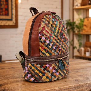 May include: A backpack featuring a multi-colored woven leather pattern in shades of red, yellow, blue, green, and purple. The bag includes solid brown leather side panels, a top handle, a front zippered pocket, and a main zippered compartment.