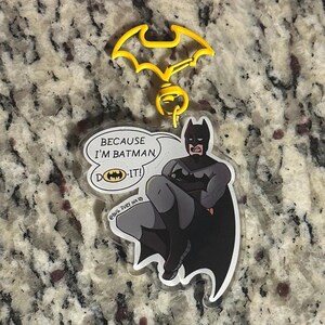 May include: A clear acrylic keychain featuring a cartoon illustration of Batman with arms crossed. A speech bubble contains the text "BECAUSE I'M BATMAN, DO-IT!" with a small bat logo. The keychain has a yellow bat-shaped plastic clasp at the top.