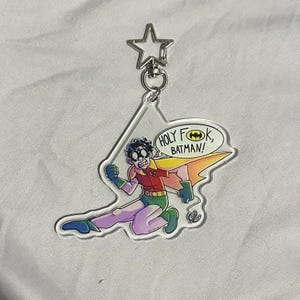 Puede incluir: Un llavero acrílico transparente con una imagen de dibujos animados de Robin de Batman. La imagen muestra a Robin con su disfraz y el texto "HOLY F*CK, BATMAN!" en un bocadillo. El llavero tiene un cierre en forma de estrella.