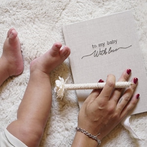 Op de afbeelding: Een wit dagboek met de tekst "to my baby with love" op de omslag. Een persoon houdt een witte pen met een bloemdetail vast en schrijft in het dagboek.