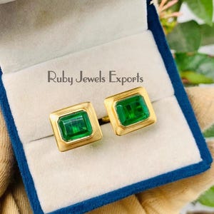 May include: A pair of gold cufflinks with emerald-cut green gemstones, presented in a blue velvet box. The cufflinks are square-shaped with a gold border. The box is open, revealing the jewelry. The text "Ruby Jewels Exports" is visible.