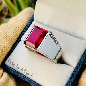 May include: A silver ring with a large, square, red gemstone and small, clear gemstones on the band. The ring is in a black box with the text "Ruby Jewels Exports" on the lid.