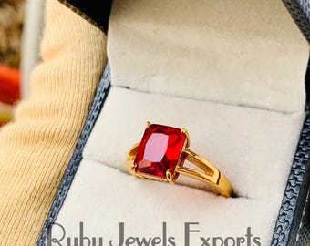 May include: A gold ring featuring a large, square-shaped red gemstone. The ring is displayed in a black box with the words "Ruby Jewels Exports" printed on it.