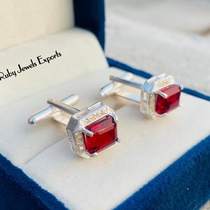 May include: Silver cufflinks with square, deep red gemstones set in a square frame, with smaller clear stones. Presented in a dark blue velvet box. The text "Ruby Jewels Exports" is visible. A stylish accessory.