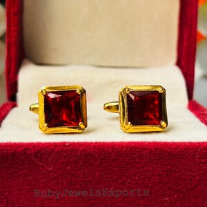 May include: Gold cufflinks with square, deep red gemstones, displayed in an open red velvet box. The cufflinks are presented on a cream-colored interior, with the brand name "Ruby Jewels Exports" visible on the box.