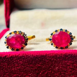 May include: Two gold rings, each featuring a large, round, red gemstone encircled by small, dark blue stones. The rings are presented in an open, red velvet jewellery box. The box displays the text "Ruby Jewels" on a white background.