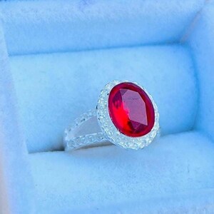 May include: A silver ring with a large oval red gemstone surrounded by smaller white gemstones. The ring is in a blue velvet ring box.
