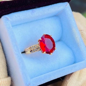 May include: A gold ring with a large, oval-cut, bright red gemstone. The ring is set in a gold band with small, clear accent stones. The ring is presented in a blue box with a burgundy velvet interior.