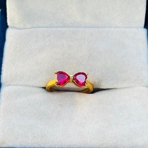 Könnte beinhalten: Ein goldener Ring mit zwei roten herzförmigen Steinen, die in Form einer Schleife angeordnet sind.
